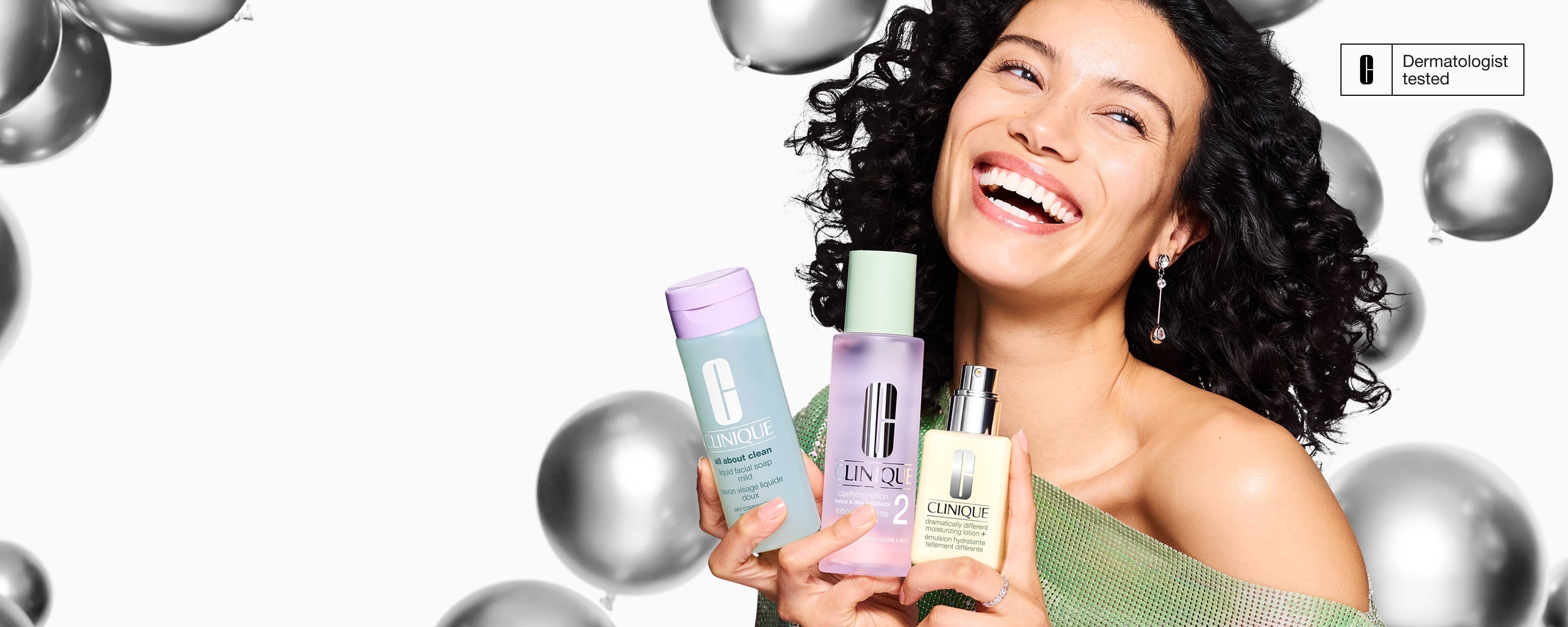 Brunette model, smiling, holding Clinique 3-Step Skincare system in front of silver balloons flying in the air