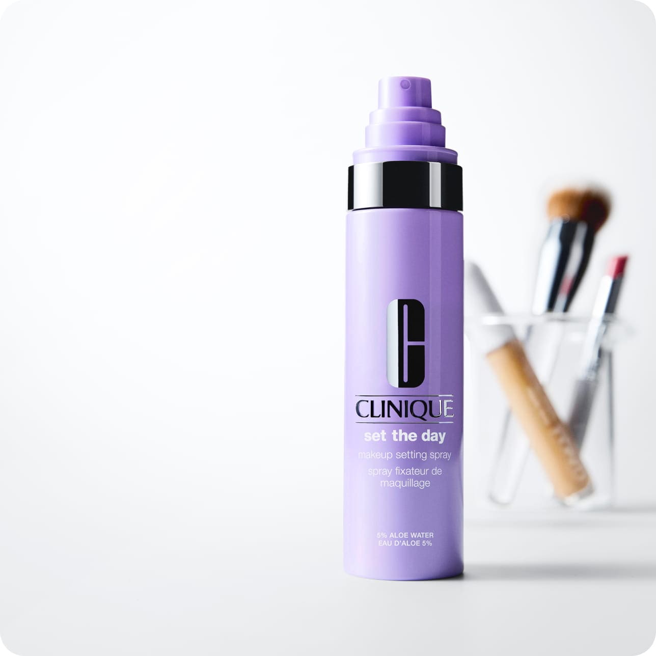 Clinique Set the Day makeup setting spray displayed beside makeup brushes in a clear holder.