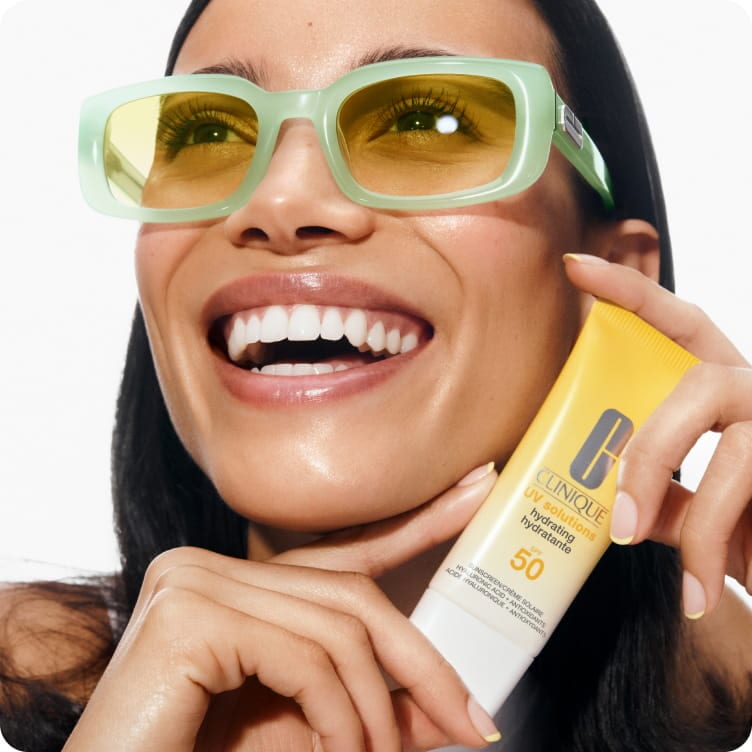 Close up of brunette model wearing green sunglasses holding hydrating sunscreen. Focusing on glowing, healthy looking skin
